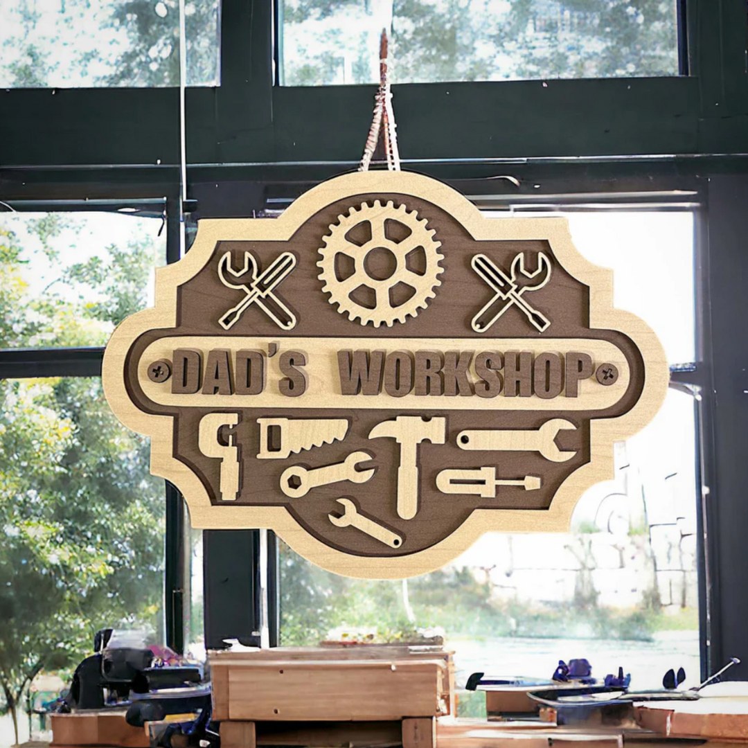 A laser cut sign with light and dark wood that says Dad's Workshop with tools and gears